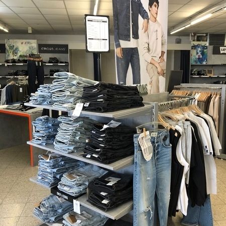 Präsentationstisch mit Jeans und Oberteilen. Auf einem zentralen Tisch liegen helle und dunkle Jeans, daneben hängen schwarze, weiße und beige Oberteile. Im Hintergrund ist Werbung für Cross Jeans zu sehen.
