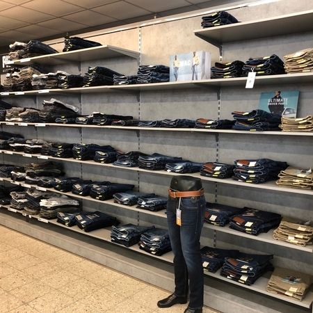 Jeansregal mit Herrenmode. Auf mehreren Regalböden liegen gefaltete Jeans in verschiedenen Farben und Schnitten. Im Vordergrund steht ein Jeansmodell auf einem schwarzen Torso mit Gürtel.