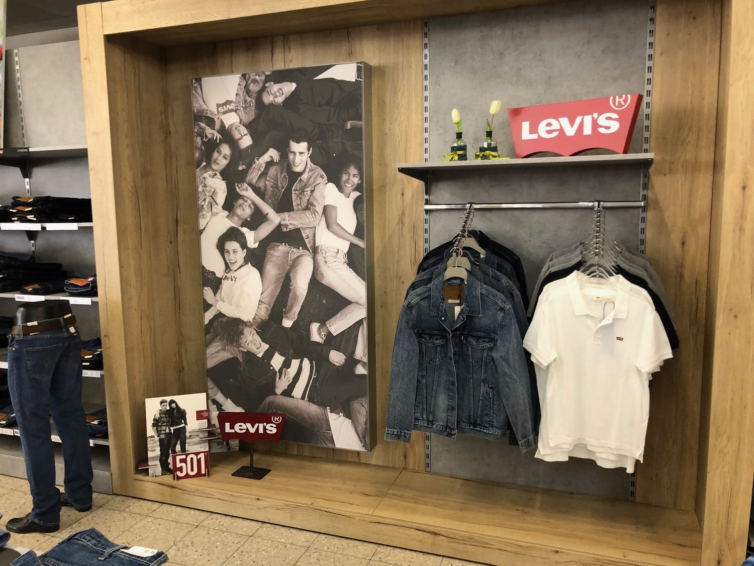 Levis Präsentation mit Herrenmode. In einem Holzrahmen sind ein großes Plakat mit Levis Motiv, eine Auswahl an Jeansjacken und Poloshirts sowie ein Levis Logo ausgestellt. Links daneben steht ein Jeansmodell auf einem Sockel.