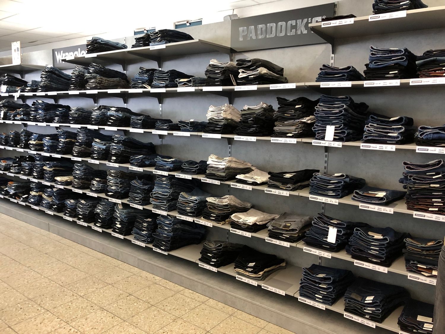 Jeansregal mit Herrenhosen. Zahlreiche gefaltete Jeans der Marken Wrangler und Paddocks liegen ordentlich sortiert auf mehreren Regalböden in einem Bekleidungsgeschäft.