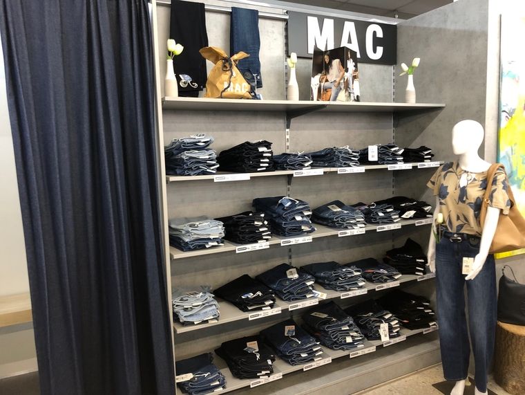 Jeansregal mit MAC Damenmode. Gefaltete Jeans liegen auf mehreren Regalebenen unter dem Markenschild MAC. Eine Schaufensterpuppe mit gemustertem Shirt und dunkler Jeans steht rechts neben dem Regal.