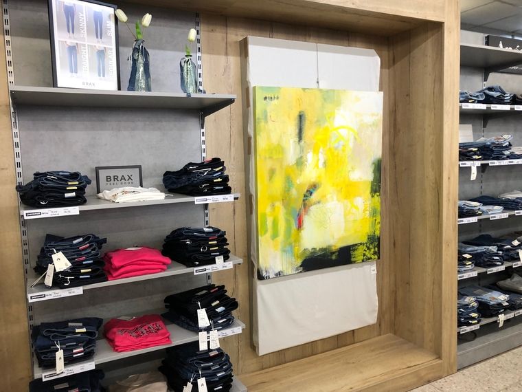 Verkaufsfläche mit BRAX Jeans und Kunstbild. Links im Bild liegen gefaltete Jeans und pinke Oberteile in einem Regal mit BRAX Logo. In der Mitte hängt ein großformatiges abstraktes Bild mit gelben und weißen Farbakzenten.