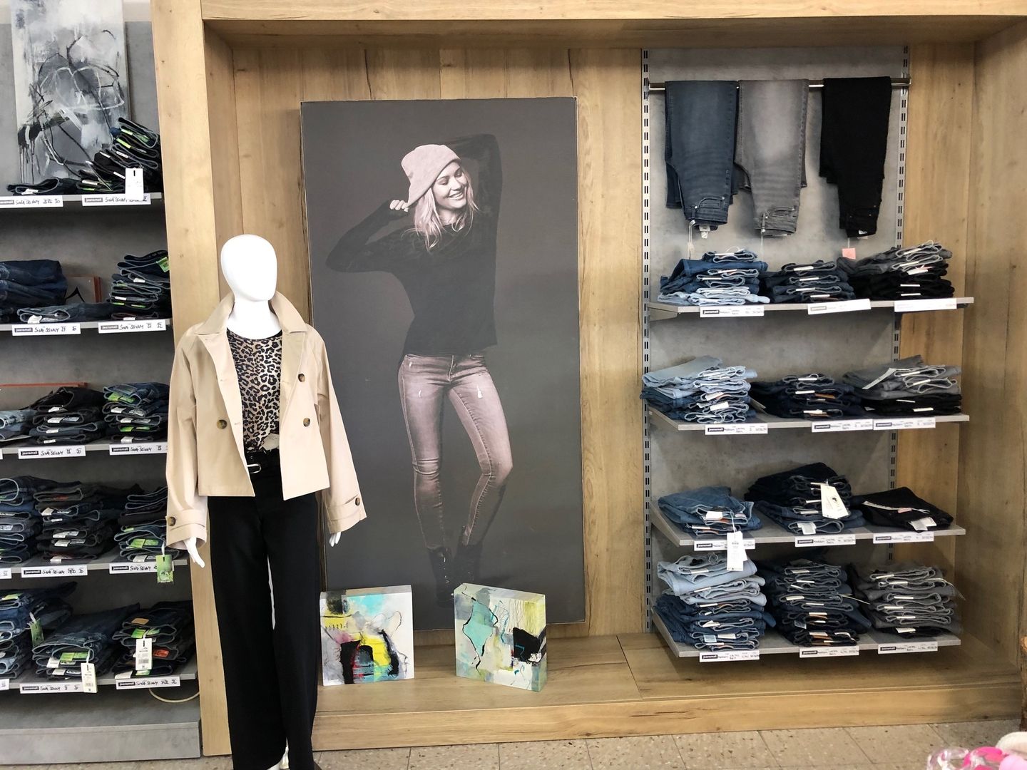 Modepräsentation mit Jeansregal und Plakat. Eine Schaufensterpuppe trägt helle Jacke und schwarze Hose. In der Mitte steht ein großformatiges Plakat mit einer lächelnden Frau in Jeans und Pullover. Rechts und links befinden sich ordentlich gefaltete Damenjeans im Regal.