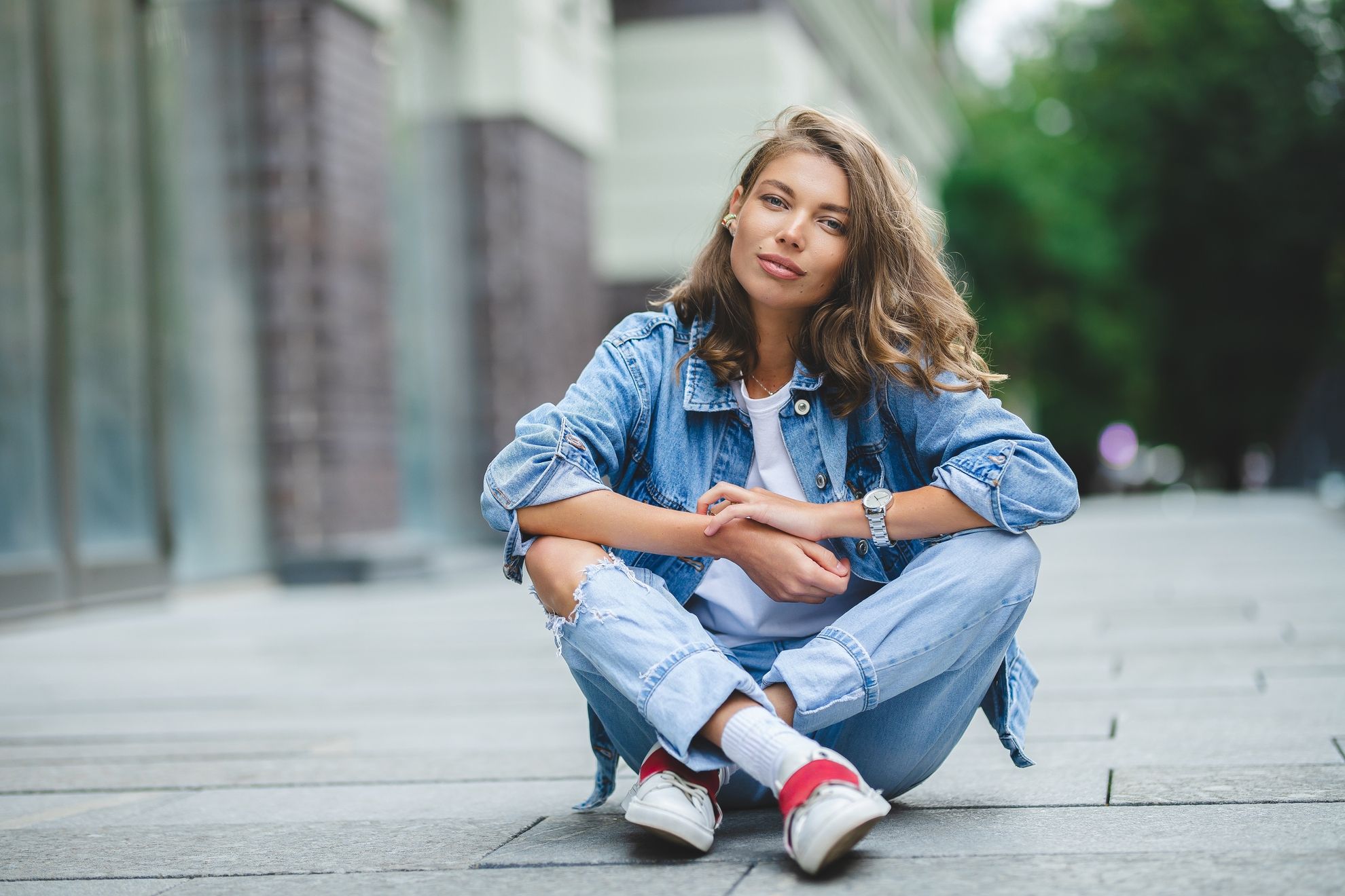 Junge Frau in Jeansoutfit sitzt auf dem Boden: Eine Frau mit langen, gewellten Haaren trägt eine Jeansjacke, ein weißes T-Shirt und hellblaue Jeans mit Rissen. Sie sitzt entspannt auf dem Gehweg in urbaner Umgebung und blickt in die Kamera.